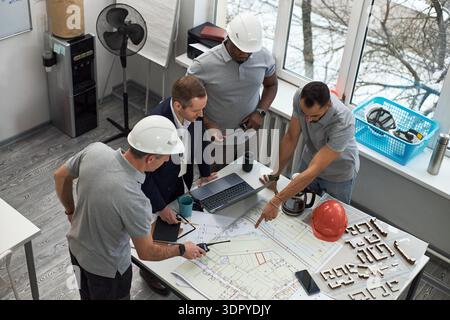Group of middle aged Caucasian man and Black men collaborating over construction blueprints in office, foreman leading discussion while pointing at plans, laptop and safety helmets visible Stock Photo
