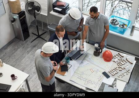 Middle aged Caucasian man in suit collaborating with two middle aged Black men and one middle aged Caucasian man wearing hard hats, reviewing blueprints and documents in office Stock Photo