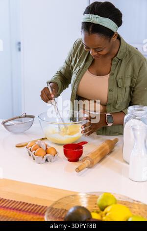 African American woman in olive-green shirt mixing batter at kitchen island with glass bowl, whisk Stock Photo