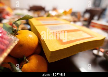 Yellow ceremonial paper stacks beside fresh oranges on table, blurred background of traditional Asian altar setup Stock Photo