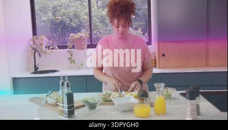 Chopping avocados on wooden cutting board at kitchen island, woman in pink tee wearing smartwatch Stock Photo