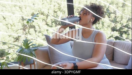 Drinking woman in gray athletic wear sitting on beige sofa at home, with metal water bottle Stock Photo