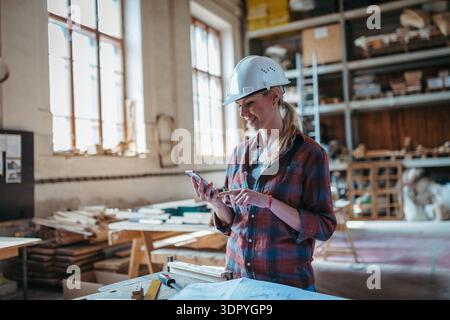 Female woodworker looking at email from client, standing in carpentry workshop. Stock Photo