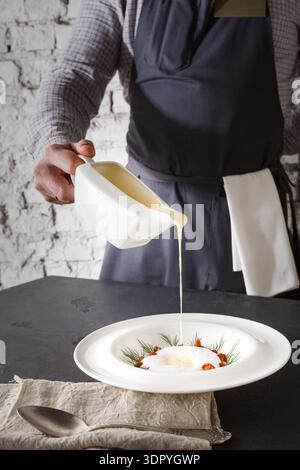 Chef Pours Sauce Over a Dish in a Kitchen Setting With Stone Wall Background Stock Photo