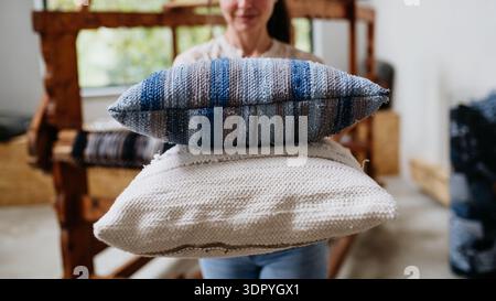 Young businesswoman next to wooden loom, holding pillows with woven covers made from recycled fabrics. Upcycling in business and circular economy. Stock Photo