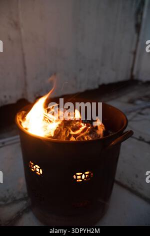 Burning coals in metal fire pit with glowing embers, outdoor evening scene near wooden wall Stock Photo