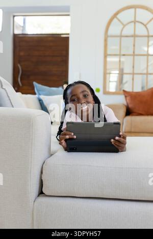 Youth female wearing pink top lying across beige sofa holding dark tablet case in living room Stock Photo