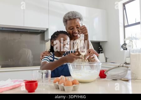 African Senior woman guiding Youth girl in apron mixing batter in kitchen with glass bowl eggs Stock Photo