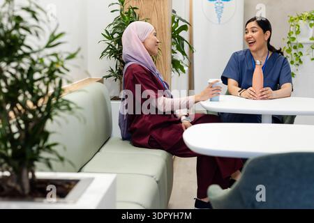 Diverse female health workers in scrubs sitting and talking in staff lounge, holding orange bottle Stock Photo