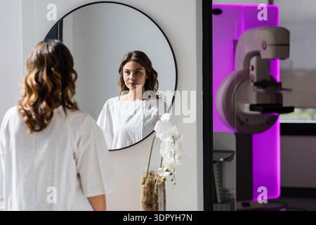 Woman in her thirties standing in medical gown, looking into round mirror near mammography machine Stock Photo