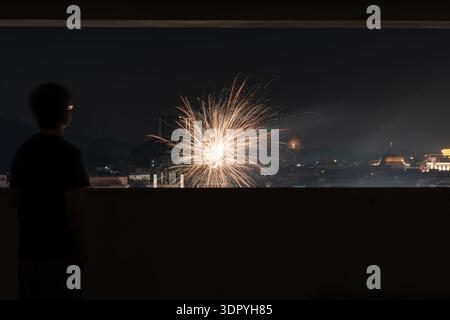 Person watching fireworks display at night from rooftop overlooking city skyline with explosion and light trails Stock Photo