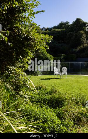 Ornate white chairs are facing each other with round table on manicured lawn near glass fence Stock Photo