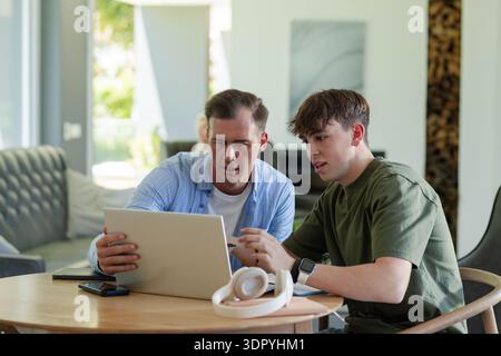 Father and teenage son using silver laptop at home, leaning, son wearing olive tee with smartwatch Stock Photo