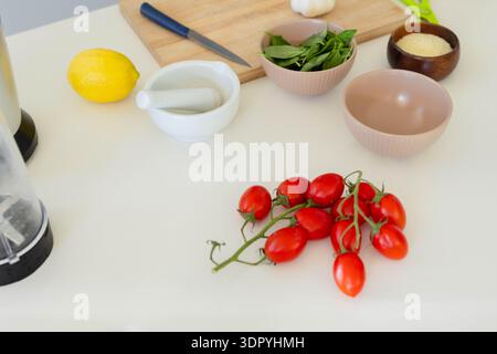 Cluster of red vine tomatoes sitting on white countertop with cutting board, knife, basil, lemon Stock Photo