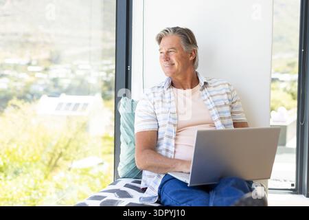 Senior man sitting on window seat using laptop, wearing striped shirt, patterned throw and cushion Stock Photo