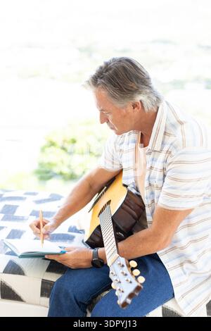 Senior man sitting on patterned bench near bright window, holding guitar and writing in notebook Stock Photo