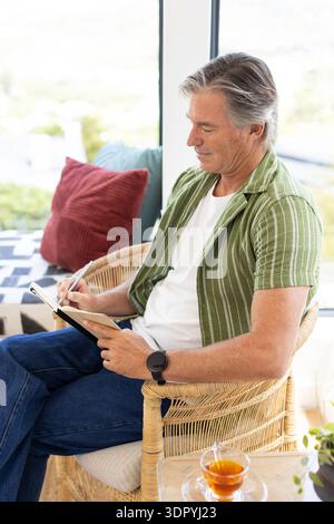 Mature man sitting in rattan armchair in sunroom writing in notebook with pen and tea Stock Photo