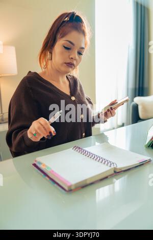 Woman standing at a home desk while checking written notes and comparing information on her smartphone. Concept of verification, focus, organization a Stock Photo