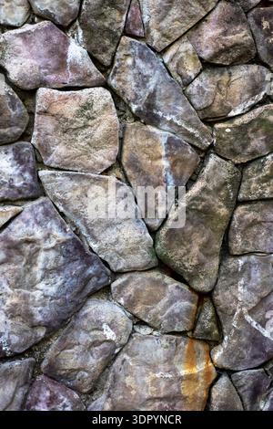 Weathered stone wall with mossy joints revealing patchwork of flagstone and slate, coarse granite faces, aged mortar Stock Photo