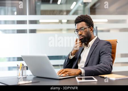 Thoughtful african american businessman is concentrating on work, using a laptop and tablet at his modern office desk, contemplating new strategies and analyzing data Stock Photo