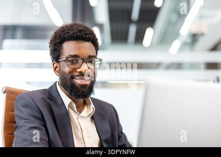 Young professional man with a friendly expression and eyeglasses sitting at a desk with a laptop, reflecting trust, corporate success, and a positive career journey Stock Photo