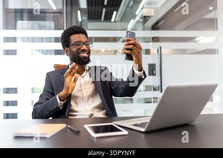 Smiling african businessman participating in a virtual meeting or a video conference from his professional office workspace, communicating remotely with colleagues or clients via a mobile phone Stock Photo