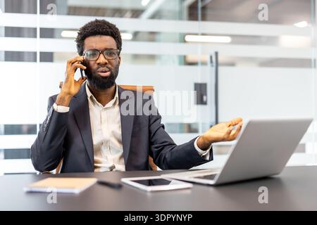 Stressed businessman talking on cell phone while sitting at office desk with laptop and tablet, expressing frustration and misunderstanding during a challenging conversation Stock Photo