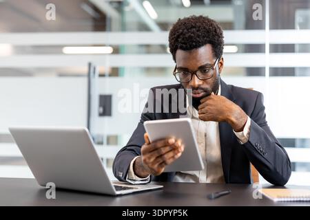 African american businessman sitting at modern office desk, wearing glasses and a formal suit, using a digital tablet while concentrating on a professional task Stock Photo
