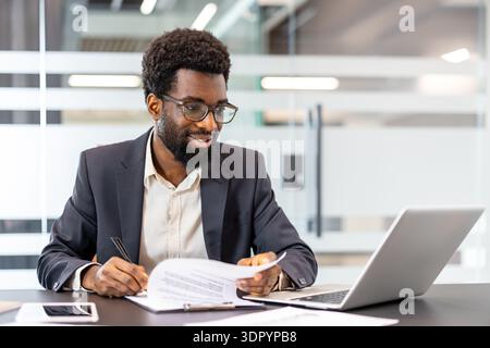 Smiling professional man in a suit working at an office desk, reviewing documents and using a laptop, embodying productive work and corporate focus in a modern workspace Stock Photo