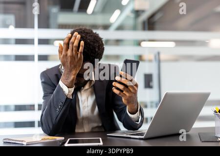 Stressed professional covering face with hands while holding a mobile phone, experiencing headache and frustration at a modern office desk with a laptop and pen holder Stock Photo