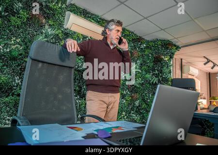 Senior professional discussing work on smartphone while standing near green wall in modern office with laptop and documents Stock Photo