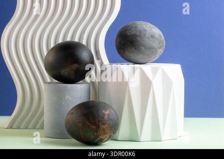 Easter eggs in black color on decorative white podiums on a blue background. Still life, copy space. Stock Photo
