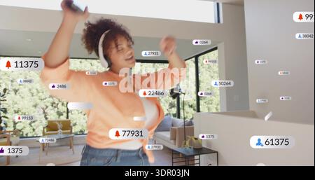 Dancing woman holding phone, wearing white headset, orange tied shirt, jeans at home showing badges Stock Photo