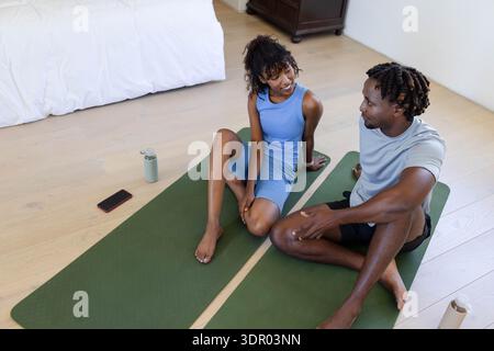 African American couple sitting on green yoga mats in bedroom in workout clothes smartphone nearby Stock Photo