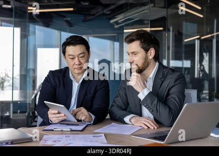 Two multi-ethnic businessmen are collaborating on a digital tablet and documents during a professional business meeting in a modern corporate office, fostering teamwork and strategic planning Stock Photo