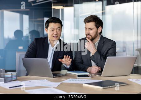 Two diverse male business professionals discussing project information while collaborating on laptops and analyzing documents during a corporate meeting in a modern office Stock Photo