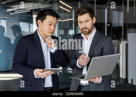 Two diverse businessmen standing in a modern office, one holding a laptop and the other a tablet, collaborating and discussing business strategy while analyzing data Stock Photo
