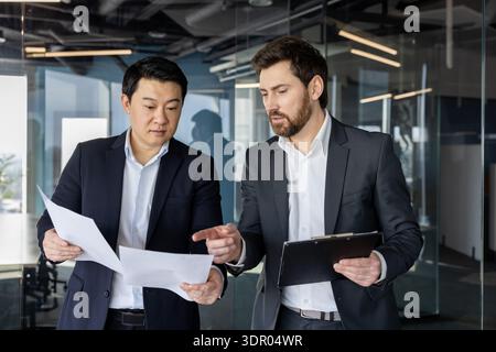 Diverse business professionals discussing documents and collaborating on a project in a modern glass office, showcasing teamwork and partnership during a business meeting Stock Photo
