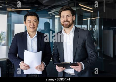 Two professional businessmen, asian and caucasian, standing in a modern glass office holding documents and smiling confidently to convey teamwork, partnership and corporate success Stock Photo