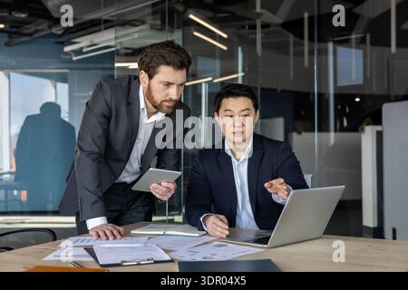 Diverse businessmen are collaborating on a project, analyzing data and charts on a laptop and tablet during a meeting in a contemporary office setting, highlighting teamwork and executive discussion Stock Photo