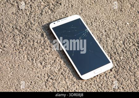 A high-angle shot of a white smartphone with a severely cracked and shattered glass screen lying on rough outdoor asphalt, symbolizing digital damage Stock Photo