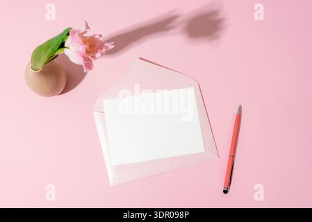 Pink envelope with blank card and pen near tulip in vase on pastel background with copy space. Mothers day concept. Top view, mockup. Stock Photo