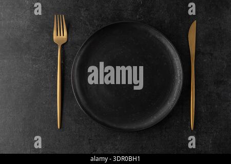 Empty grunge dark grey craftmade ceramic plate with golden cutlery ( fork, knife ) on black stone board background. Retro style table serving matte Stock Photo