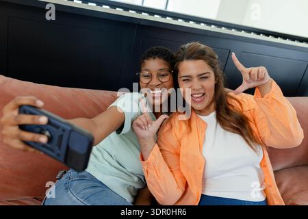 Diverse female friends wearing glasses sitting on peach-colored couch taking selfie with smartphone Stock Photo