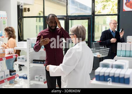 African American male customer consults senior pharmacist about antibiotics and headache medication at pharmacy aisle. Sick man explains symptoms, seeking professional guidance at local dispensary. Stock Photo