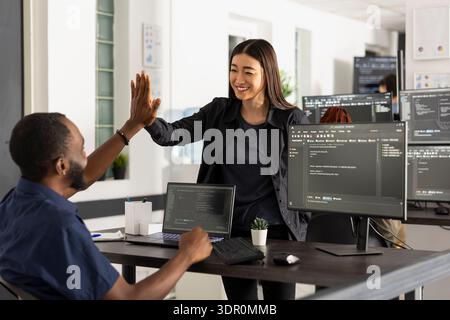 Colleagues celebrate coding project completion, cooperation and teamwork, capturing positive, professional workspace. African american and asian programmers smiling and gesturing in startup office. Stock Photo