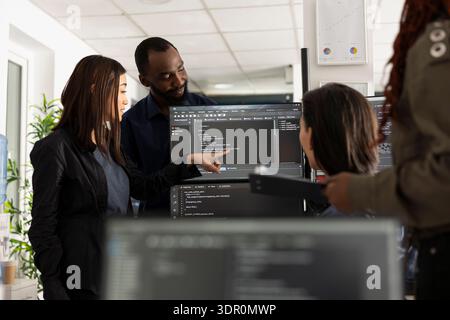 African american and asian coworkers collaborate at desk, comparing code and planning website updates on desktop pc. Professional team reviews app functionality with focus and innovation mindset. Stock Photo