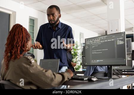 Confused man speaks with colleague, discussing updates to website functionality. Male and female developers collaborate at computer, working to improve coding scripts and system performance. Stock Photo