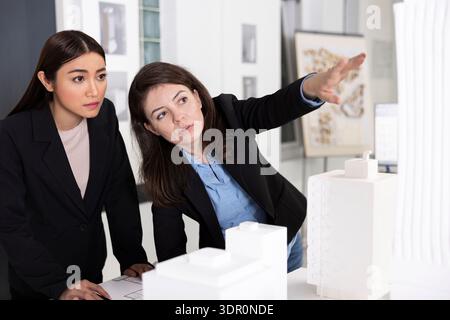 Architects analyze detailed architectural model while discussing structural ideas and project layout. Professional women show teamwork, precision and creative problem solving in modern design office. Stock Photo