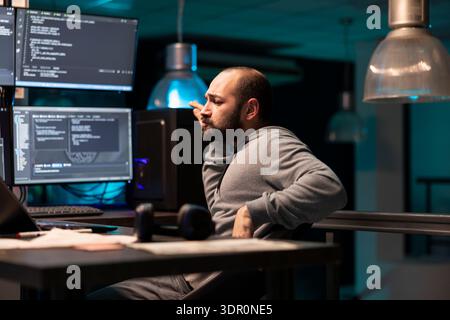 Male coder sitting and dancing with satisfaction after solving complex programming issue. Tech professional enjoys well earned victory in modern office lit by glowing computer monitors. Stock Photo